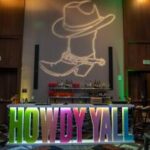 Event bar with large illuminated “Howdy Y’all” sign and cowboy boot projection—welcoming western touch designed by a North Texas DMC.