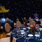 Formal dining tables set for a gala with navy linens, floral centerpieces, and a starlit backdrop—meticulously executed by Texas corporate event planners.